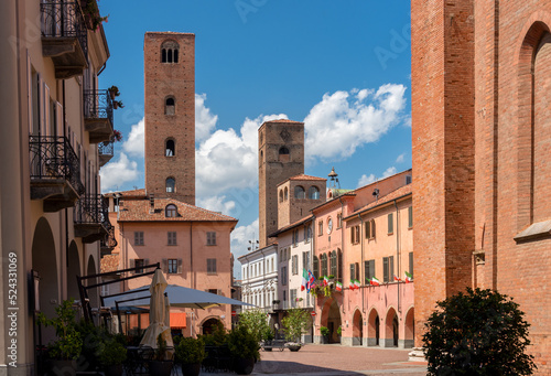 View of Duomo square with the town hall among old houses and medieval towers under beautiful sky in Alba, Langhe, Piedmont, Italy.