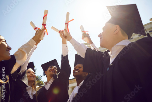 Happy diverse college or university students having fun at graduation. Joyful multiracial graduates standing together and holding up paper diploma scrolls with red ribbons. Education, success concept