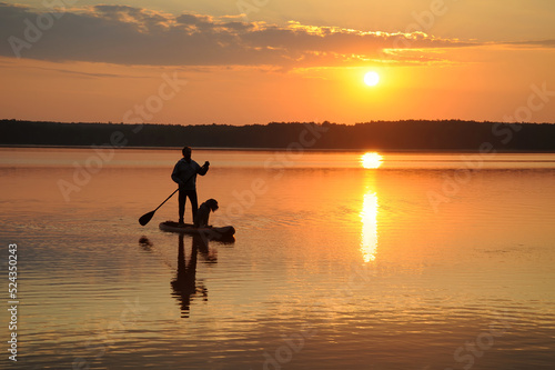 Silhouettes of a man with a dog paddling in lake water on a SUP board at sunset. Vacation, tourism, active lifestyle, hobbies.
