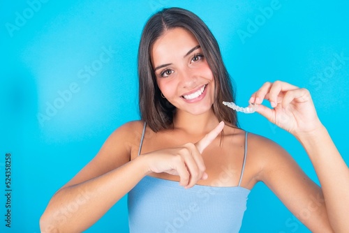beautiful brunette woman wearing blue tank top over blue background holding an invisible aligner and pointing at it. Dental healthcare and confidence concept.