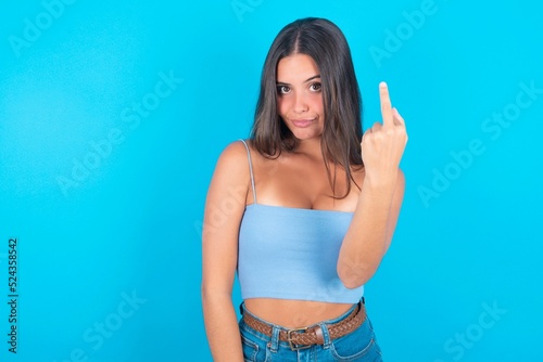 beautiful brunette woman wearing blue tank top over blue background shows middle finger bad sign asks not to bother. Provocation and rude attitude.