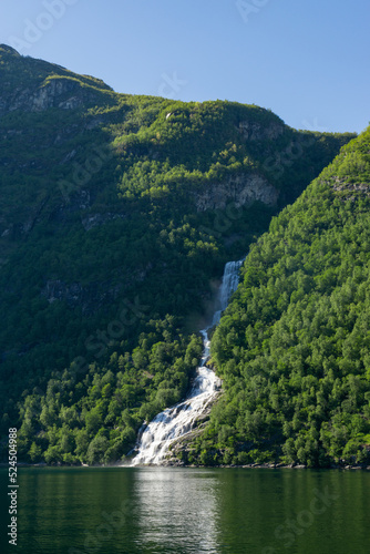 Waterfall in the Geiranger fjord surrounded by green trees and high mountains in Norway.
