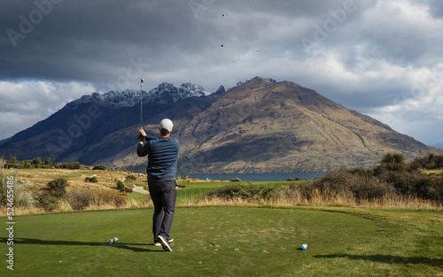 Queenstown, Otago, New Zealand - 04.24.21:Young male golfer hits tee shot on the iconic 11th hole at Jack's Point. Beautiful views of Lake Wakatipu and snow topped mountain ranges beyond.