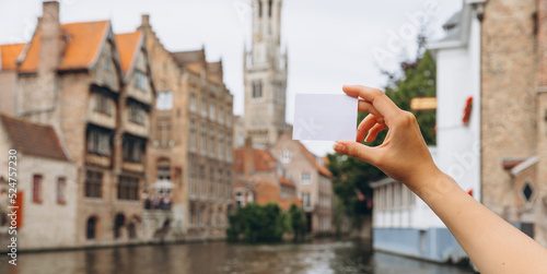 Female hand holding a ticket or business card with copy space on the background of a canal and medieval houses in Bruges, Belgium