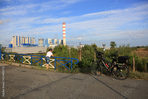 Mężczyzna oparty na poręczy wiaduktu kolejowego, z rowerem turystycznym, elektrownia.