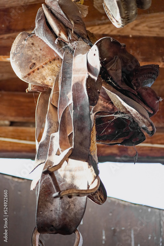 This is a digitally enhanced image of an antique horse saddle hanging from the rafters of a barn. A transom window provides enough natural light to see the cracked leather and brown and red colors.