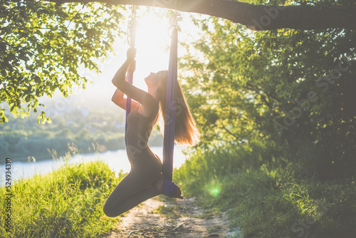 A young female gymnast is engaged in aerial yoga, using a combination of traditional yoga poses, pilates and dance using a hammock at sunset in nature. Healthy lifestyle.