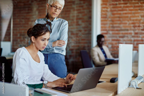 Young businesswoman working on laptop while senior executive is supervising her in office.