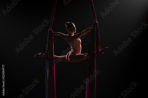 Flexible aerial gymnast performs a twine on red air silk. View from back of female acrobat is balancing on black background with backlight. Young woman performs tricks at height on a red silk fabric.