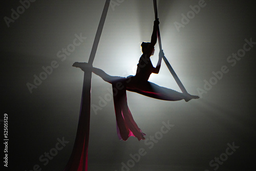 Dark silhouette of a young woman equilibrist demonstrating twine, hovering on an airy silk at height. An acrobat performs gymnastic tricks in dark with center light. Balancing on silk ribbon.