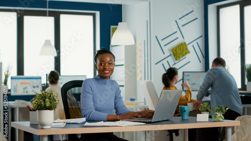 Portrait of smiling african american woman working on profit margin and sale statistics using laptop sitting at desk in busy startup office. Casual business employee happy with job result.