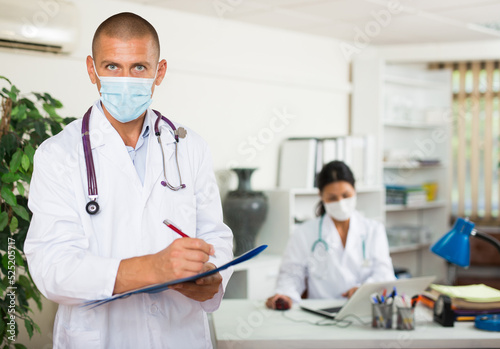 Focused therapist in white coat and protective face mask standing in clinic, filling clipboard with medical records