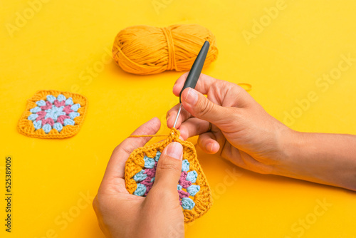 Closeup of woman's hands with a hook inserted in crochet element with the backgound of yellow table, yellow skein of yarn and finished granny square element