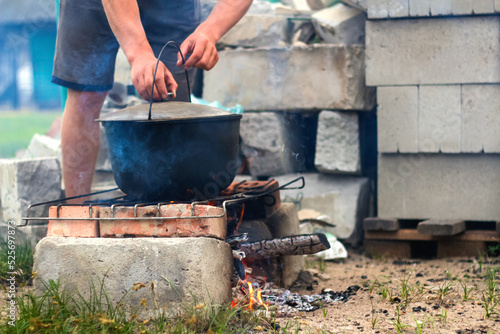 Defocus man cooking fish soup in the iron bowler over a campfire. Fish soup boils in cauldron at the stake. Soup in a pot in the fire. Camp at day. Nature green grass background. Out of focus