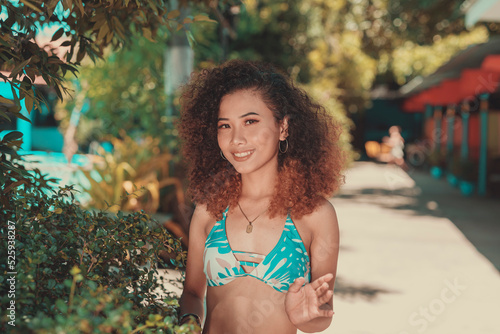 Smiling young adult in a resort hotel wearing her bikini and a newly permed hair poses near plants.