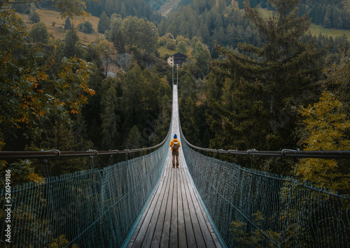 Young traveler man dressed in yellow jacket crosses hiking on an impressive wooden and metal bridge in the village of Goms in the Swiss alps