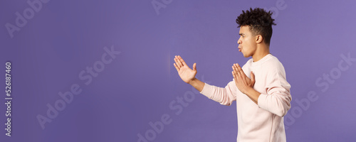 Profile portrait of young hispanic guy with dreads acting like he is ninja or martial arts fighter, practice his kung-fu or taekwondo skills, standing purple background