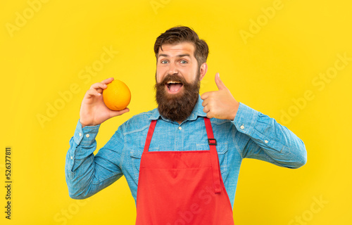 Happy man in apron giving thumb to fresh orange citrus fruit yellow background, fruiterer