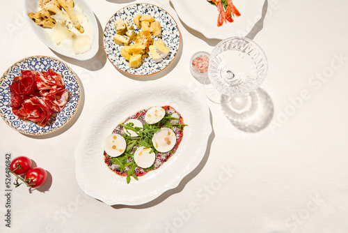 Italian antipasti beef carpaccio with parmesan, ruccola and mushrooms on white table. Carpaccio with raw meat and cheese in italian style with sunny shadows. Italian menu. Aesthetic food composition.