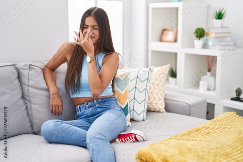 Young brunette woman sitting on the sofa at home smelling something stinky and disgusting, intolerable smell, holding breath with fingers on nose. bad smell