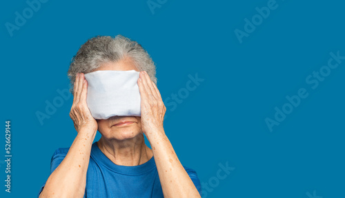 Senior woman using hot compress gel on eyes while standing on a blue background