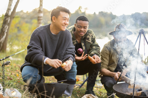 Three multiracial friends cooking Ukha soup in cauldron on bonfire during a picnic. Men fishing and having fun together on lake coast. Leisure, weekend and vacation in nature