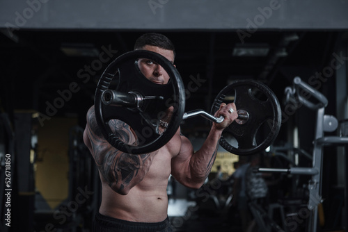 Focused muscular shirtless man lifting heavy barbell, doing biceps workout