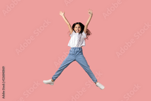 Full length portrait of charming beautiful little girl wearing white T-shirt jumping in air and raised arms, expressing excitement, having fun. Indoor studio shot isolated on pink background.