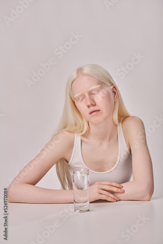 Young albino girl with white long hair sitting at table with glass of water on white background