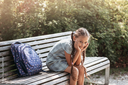 Back to school. A cute little schoolgirl in a dress with pigtails and large blue backpackis sitting on a bench in the school yard . A little girl is going to the first grade.