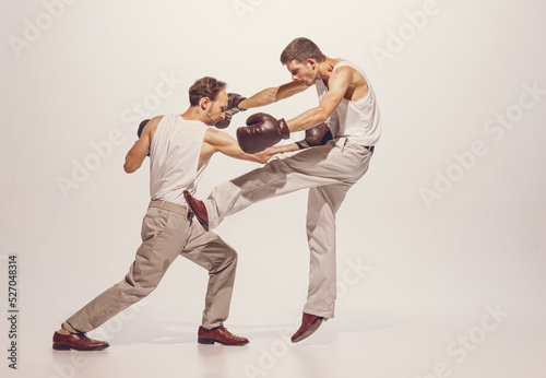 Portrait of two men playing, boxing in gloves isolated over grey studio background. Dynamic image of fight