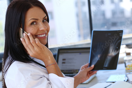Young, professional, confident female doctor with stethoscope holding X-ray and talking on phone at doctor's office