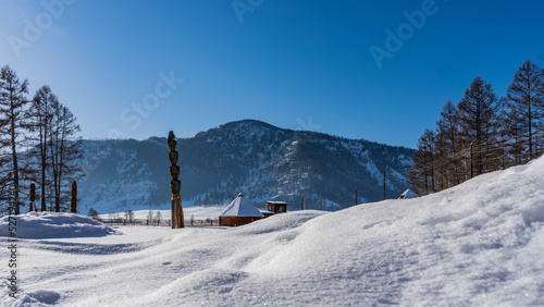 In the snow-covered valley there are traditional Altai houses - ail with a pyramidal roof. Pure white snow in the foreground. A mountain against a clear blue sky. Altai
