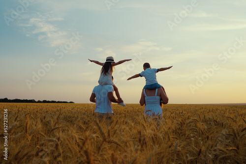 Mom and dad carrying kids on shoulders while walking in wheat field at sunset, boy and girl spreading arms as if flying, blue sky on background. Happy family together