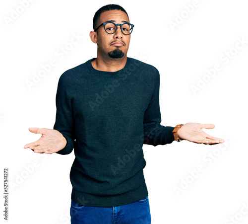 Young african american man wearing casual clothes and glasses clueless and confused expression with arms and hands raised. doubt concept.