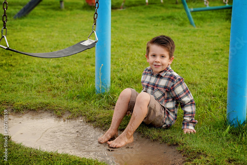 Niño feliz sentado mojando sus pies en un charco de agua con lodo y tierra disfrutando y jugando emocionado en el parque al aire libre en un hermoso día 