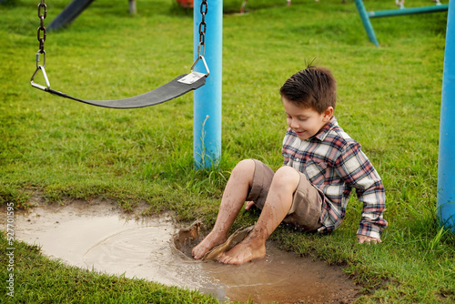 Niño feliz sentado mojando sus pies en un charco de agua con lodo y tierra disfrutando y jugando emocionado en el parque al aire libre en un hermoso día 