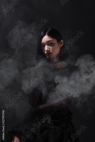 brunette young asian woman in gothic outfit and gloves on grey with smoke.