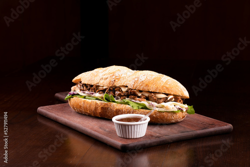 fast food beef brisket sandwich with arugula and coleslaw salad on baguette bread on wooden board with barbecue sauce dark background