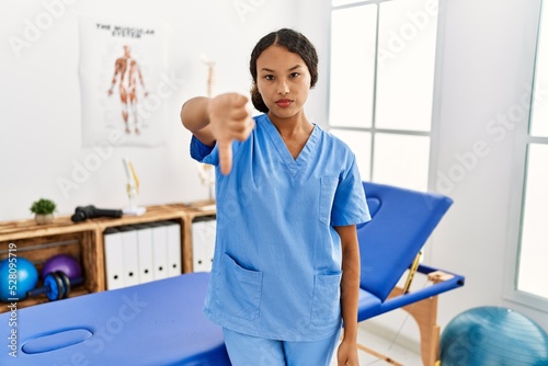 Beautiful hispanic physiotherapist woman at pain recovery clinic looking unhappy and angry showing rejection and negative with thumbs down gesture. bad expression.