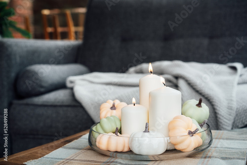 Autumn, fall cozy mood decor composition for hygge home. Decorative pumpkins and white burning candles on the glass tray on the coffee table in the living room with sofa. Selective focus