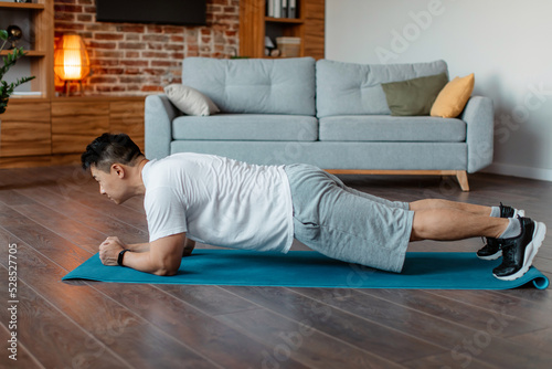 Domestic sports concept. Asian man doing plank exercises at home, training in living room on sports mat, side view