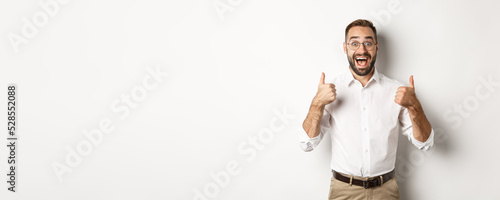 Excited man in glasses showing thumbs up and looking amazed, agree and approve something great, recommending product, standing over white background