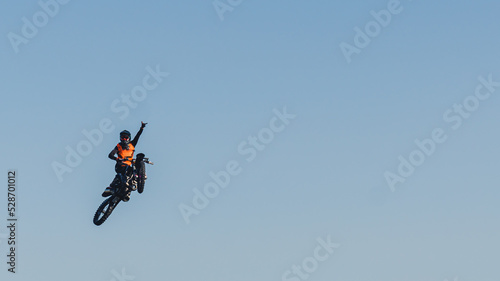 Adrenaline boost in extreme sports. Brave biker jumping extremely high on his black motorbike and waving with one hand. Blue sky as a background. High quality photo