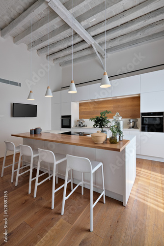interior view of a modern white kitchen, in the foreground the island kitchen with stools, the lighting is entrusted to four pendant lamps, the floor and ceiling are made of wood