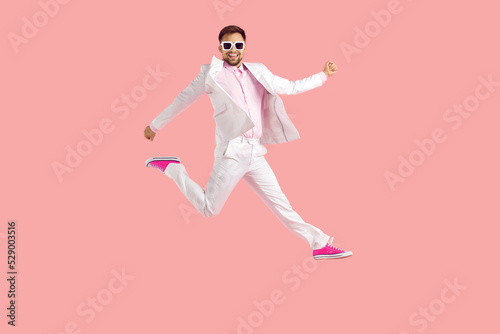 Funny confident young man having fun in the studio. Full body shot of happy excited joyful guy wearing white suit, eyeglasses and trainer shoes jumping high in the air isolated on pink background