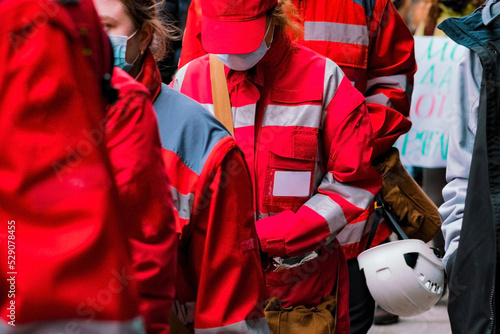 A crowd of emergency medic assistants in red uniforms with helmets stands outdoors. Paramedic. Patient. Person. Profession. Red. Rescue. Rescuer. Safety. Sick. Street Girls. Uniform. Female