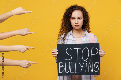 Young sad depressed black teen girl student she in casual clothes backpack bag hold card sign with stop bullying title text isolated on plain yellow background High school university college concept