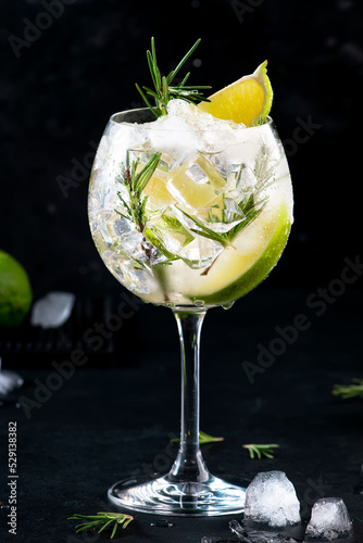Gin tonic lime alcoholic cocktail drink with dry gin, rosemary, tonic and ice in big wine glass. Black bar counter background, steel bar tools