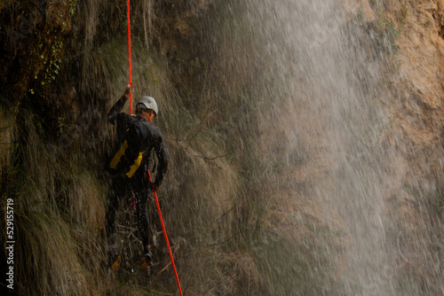 Man wearing helmet and wetsuit rappelling in the middle of a waterfall